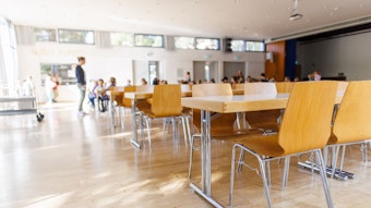 Stühle und Tische für Erwachsene stehen in einer zur Schulmensa umfunktionierten Aula einer Grundschule während im Hintergrund eine Lehrerin und Schüler zu sehen sind.