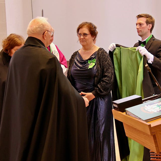 Inge Hartmann steht vor ihrem Vorgesetzten und kriegt einen grün-schwarzen Mantel umgelegt.