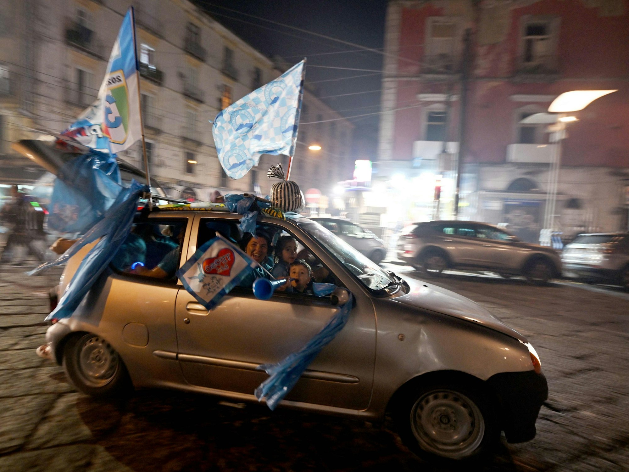 Fans von Napoli fahren in einem Auto durch die Stadt. Aus den Fenstern schwenken sie Fahnen und Schals.