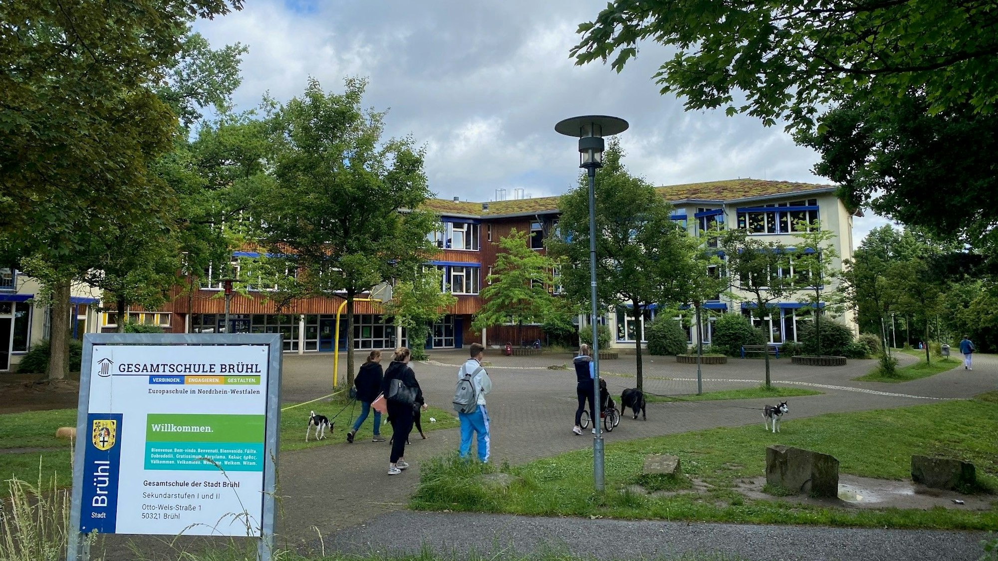 Blick auf den Schulhof der Gesamtschule. Die Gebäude werden zum Teil von grünen Bäumen verdeckt. Zwei Schüler und zwei Spaziergänger mit ihren Hunden laufen vorbei.