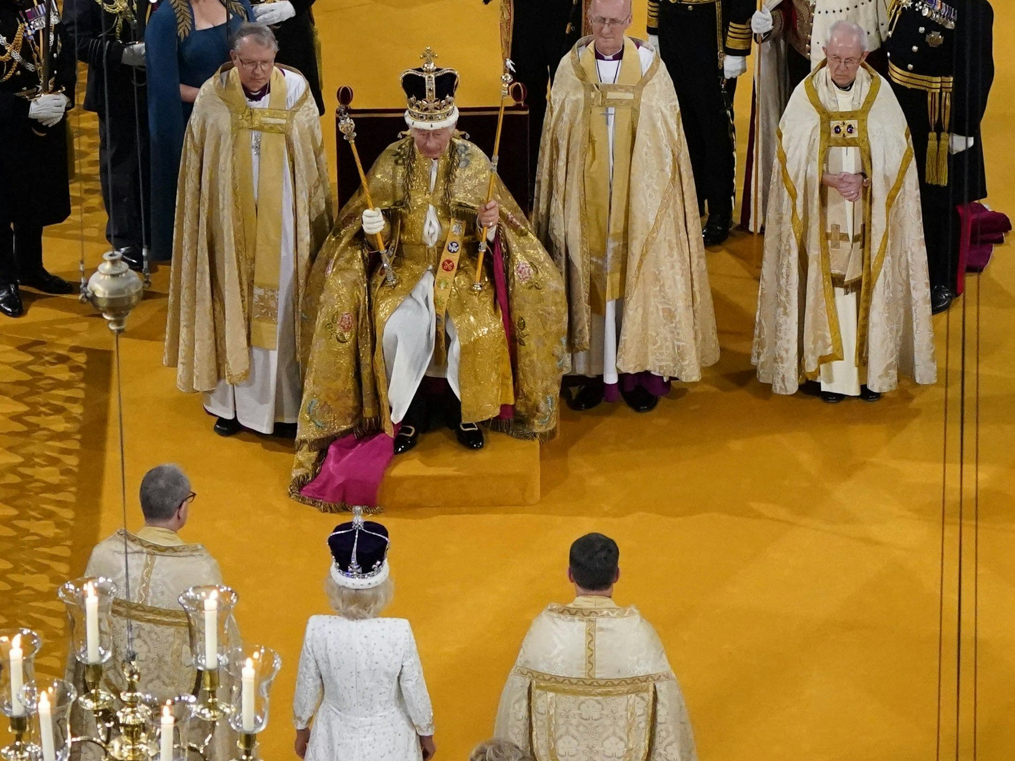 König Charles III. sitzt während der Krönungszeremonie mit der Edwardskrone in der Westminster Abbey in einem Krönungsgewand auf dem Thron, in den Händen hält er zwei Zepter, während Königin Camilla ihm gegenüber die Krone von Queen Mary trägt.