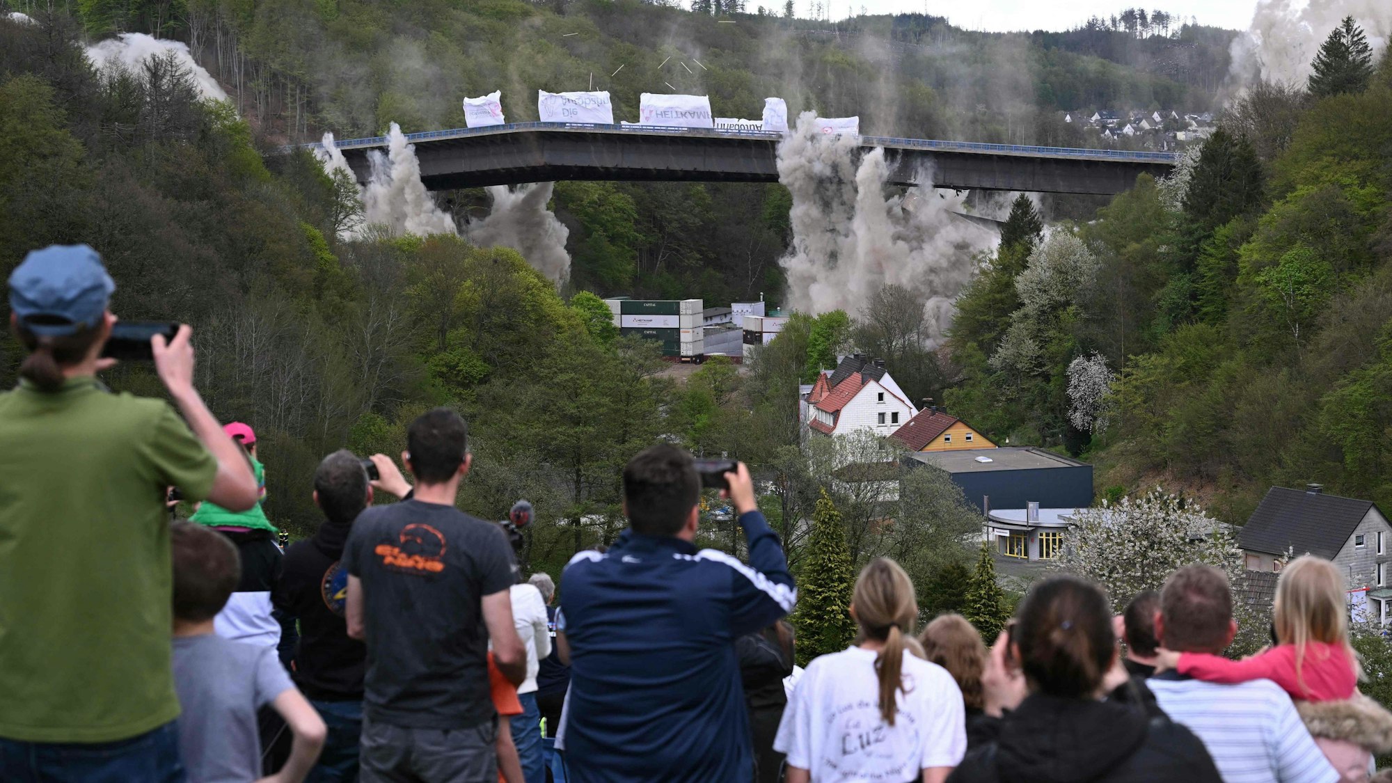 People take pictures of the Rahmede highway bridge during its blasting on May 7, 2023 on the A45 motorway, near Luedenscheid, western Germany. - During a routine inspection of the 453-metre-long Rahmede viaduct in December 2021, inspectors discovered deformations in the steel wall that could affect the bridge's load-bearing capacity. According to the motorway company, a new construction will take at least five years. (Photo by INA FASSBENDER / AFP)