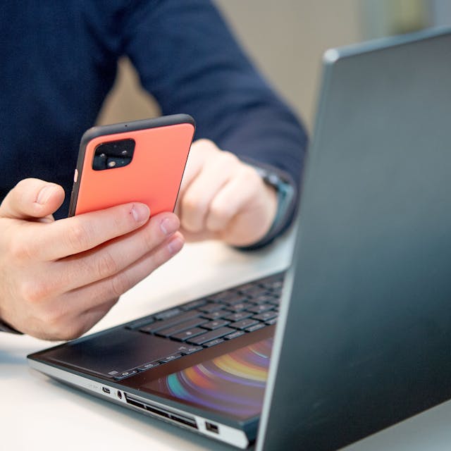 Eine Person sitzt am Schreibtisch vor einem Laptop und hält dazu das Smartphone in der Hand.