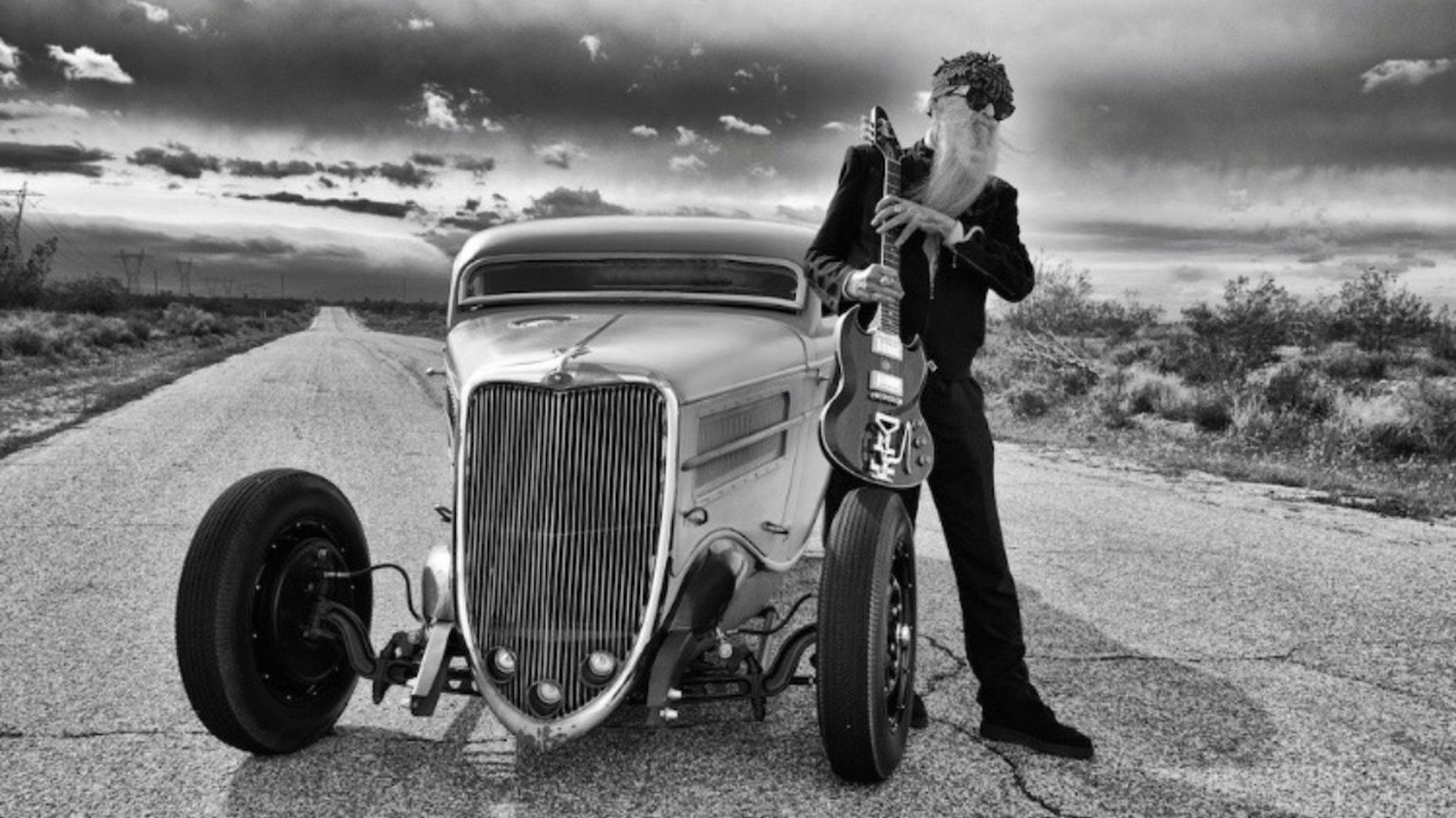 ZZ-Top-Gitarrist Billy F. Gibbons steht neben seinem Hot-Rod-Auto auf einer Wüstenstraße in den USA. In den Händen hält er seine E-Gitarre, aufgestützt auf einem Vordereifen des Wagens.