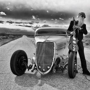 ZZ-Top-Gitarrist Billy F. Gibbons steht neben seinem Hot-Rod-Auto auf einer Wüstenstraße in den USA. In den Händen hält er seine E-Gitarre, aufgestützt auf einem Vordereifen des Wagens.&nbsp;