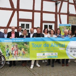 Die Vertreter der beteiligten Kreise und Kommunen halten vor einem Fachwerkgebäude in Antweiler ein Banner, mit dem für die „Tour de Ahrtal“&nbsp;geworben wird.