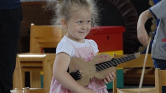 Ein kleines Mädchen mit rosafarbenem Kleid hält eine Geige aus Pappe unterm Arm.