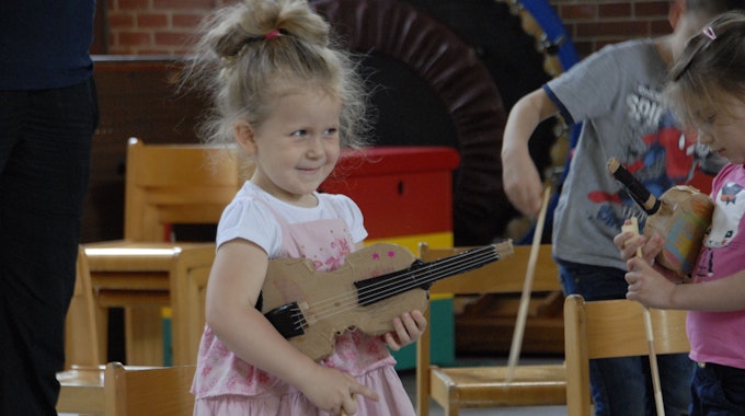 Ein kleines Mädchen mit rosafarbenem Kleid hält eine Geige aus Pappe unterm Arm.