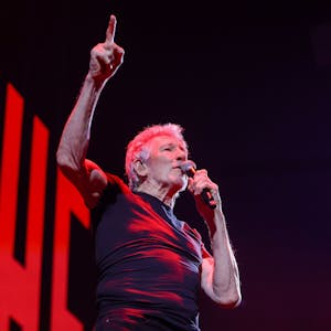 Roger Waters in der Lanxess Arena, er hält sich ein Mikro an den Mund, während er mit der rechten Hand aufzeigt.