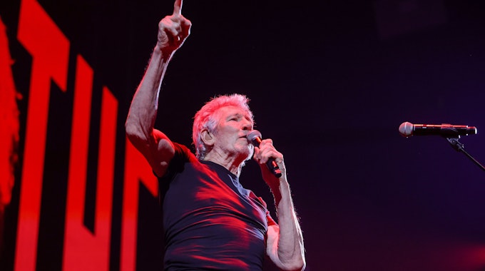 Roger Waters in der Lanxess Arena, er hält sich ein Mikro an den Mund, während er mit der rechten Hand aufzeigt.