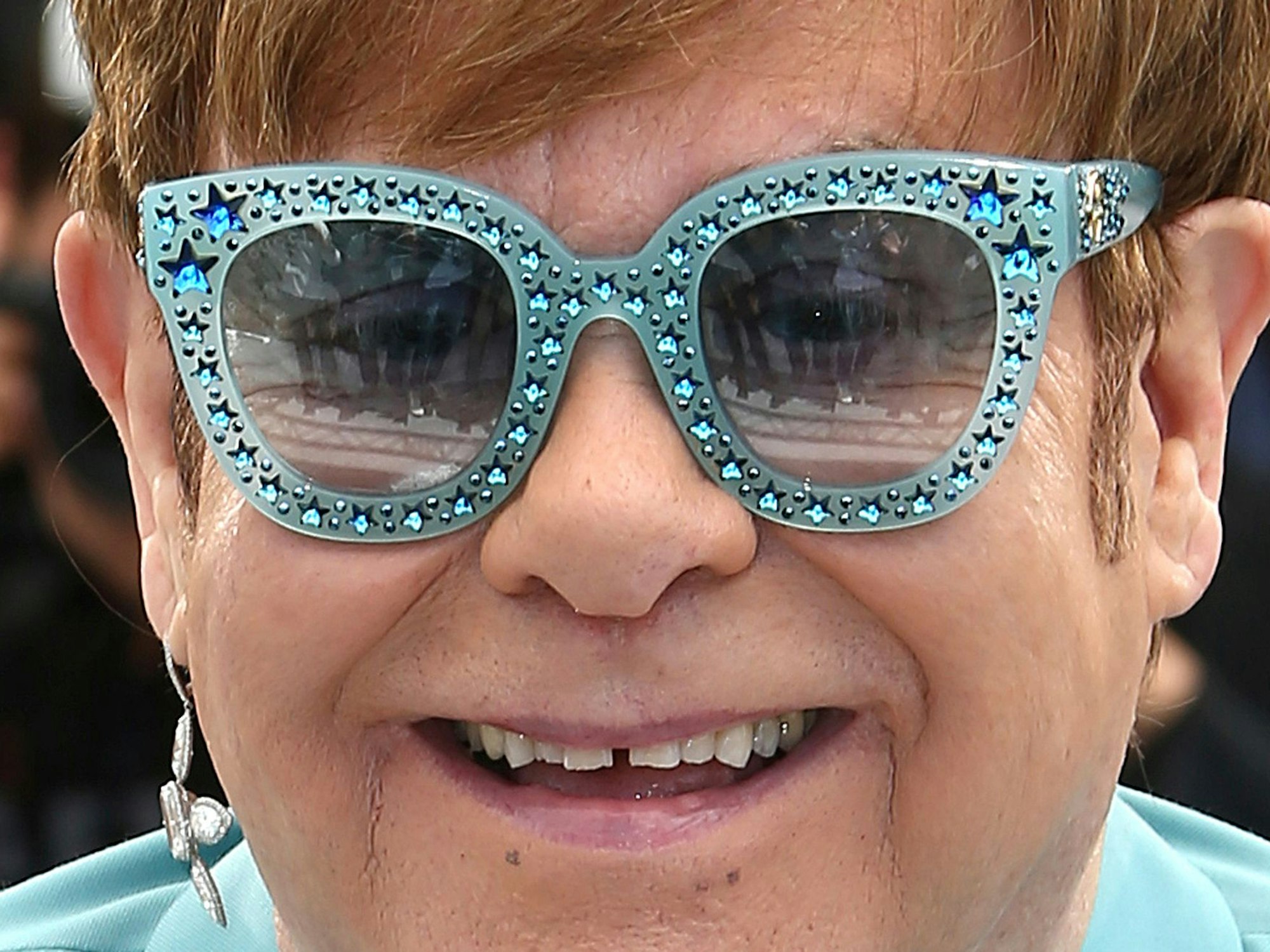 Elton John trägt eine blaue Sonnebrille und lacht