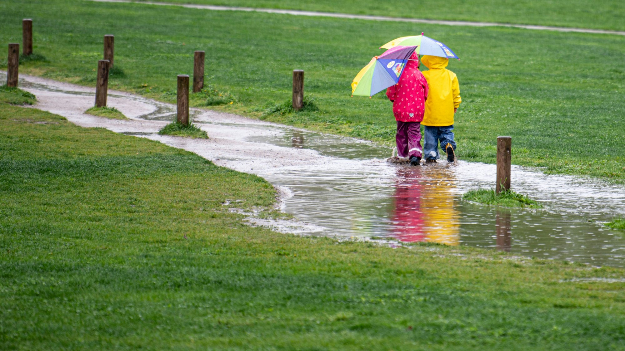 Zwei Kinder laufen in unserem Symbolbild einem Park durch eine große Pfütze. Im Westen Deutschlands lässt Starkregen die Flüsse steigen.