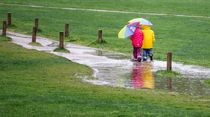 Zwei Kinder laufen in unserem Symbolbild einem Park durch eine große Pfütze. Im Westen Deutschlands lässt Starkregen die Flüsse steigen.