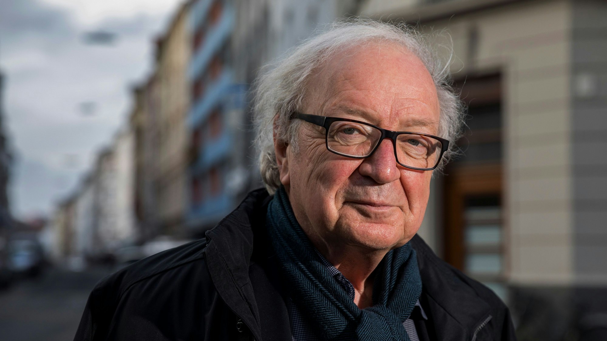 Der Journalist und Filmemacher Osman Okkan mit Brille, Hemd, Schal und Jacke und einer Kölner Straße im Hintergrund.
