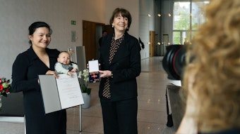 Elizaveta Khan hält ihre Urkunde in die Luft und ihr Baby im Arm, Henriette Reker hält das Bundesverdienstkreuz.