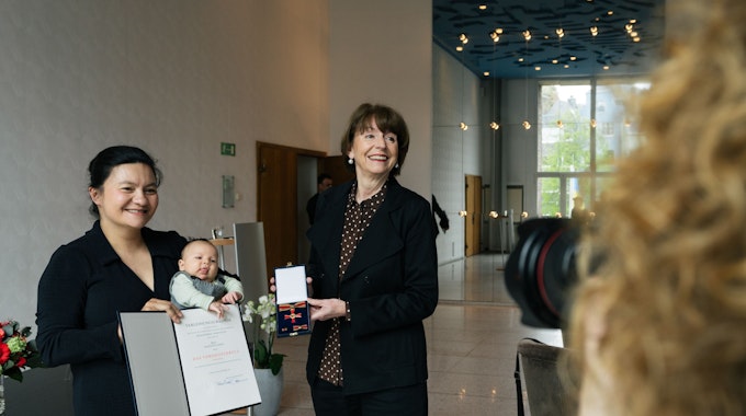 Elizaveta Khan hält ihre Urkunde in die Luft und ihr Baby im Arm, Henriette Reker hält das Bundesverdienstkreuz.