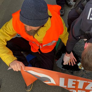 Ein Klima-Aktivist sitzt auf einer Straße in Köln, nachdem die Letzte Generation in köln den morgendlichen Berufsverkehr stellenweise lahmlegten. Er hat sich an der Straße festgeklebt und hält ein Banner der Letzten Generation. Ein Polizist versucht, seine Hand von der Straße zu lösen.&nbsp;