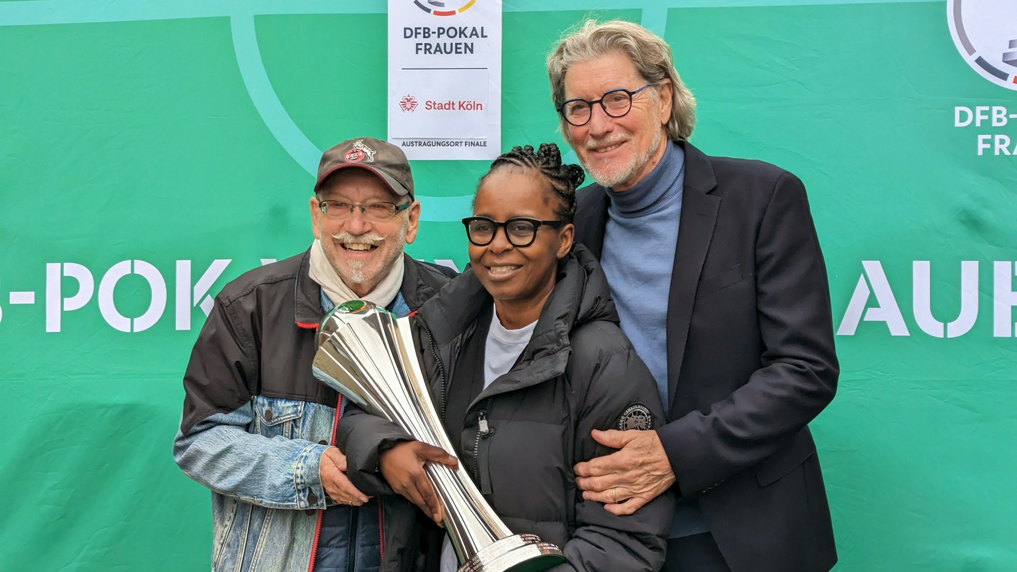 Janus Fröhlich, Shary Reeves und Toni Schumacher stehen zusammen vor einem grünen Hintergrund. Shary Reeves hat den Pokal in der Hand.
