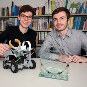 Zwei Schüler sitzen an einem Tisch, vor ihnen stehen ein kleiner Roboter und eine Siegertrophäe für einen Roboter-Wettbewerb auf dem Tisch.