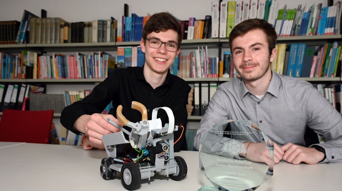 Zwei Schüler sitzen an einem Tisch, vor ihnen stehen ein kleiner Roboter und eine Siegertrophäe für einen Roboter-Wettbewerb auf dem Tisch.