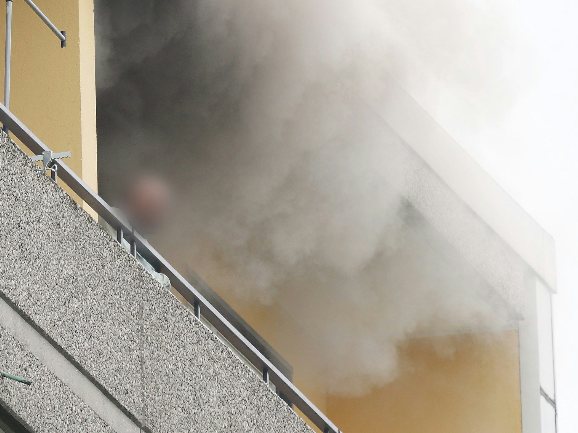 Eine Person steht auf dem Balkon von einem Hochhaus, aus dem Rauch quillt.