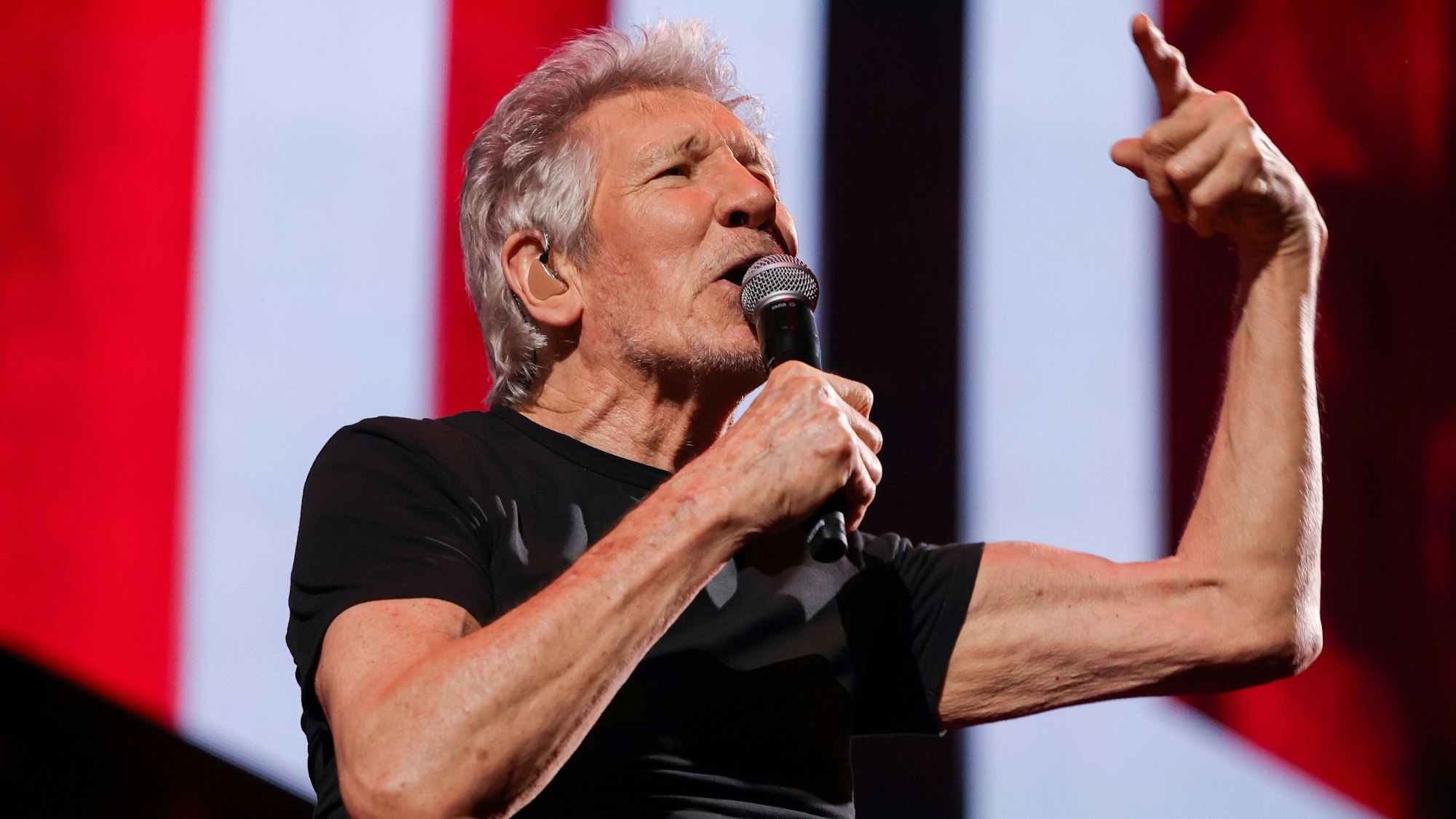 Roger Waters, in ein schwarzes T-Shirt gekleidet, singt in ein Mikrofon. Sein linker Arm ist erhoben, der Zeigefinger der linken Hand ausgestreckt. Die Bühne im Bildhintergrund schimmert rot und weiß.