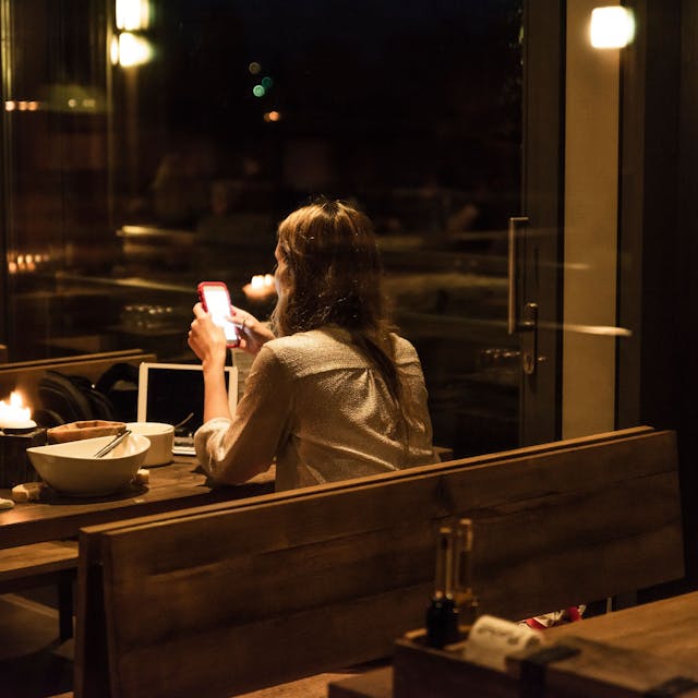 Eine Frau sitzt in einem relativ dunklen und weitestgehend leeren Restaurant an einem Tisch mit leerem Teller und schaut auf ihr Smartphone.