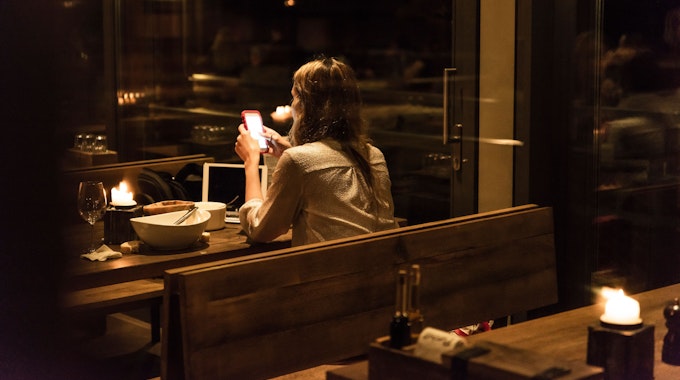 Eine Frau sitzt in einem relativ dunklen und weitestgehend leeren Restaurant an einem Tisch mit leerem Teller und schaut auf ihr Smartphone.