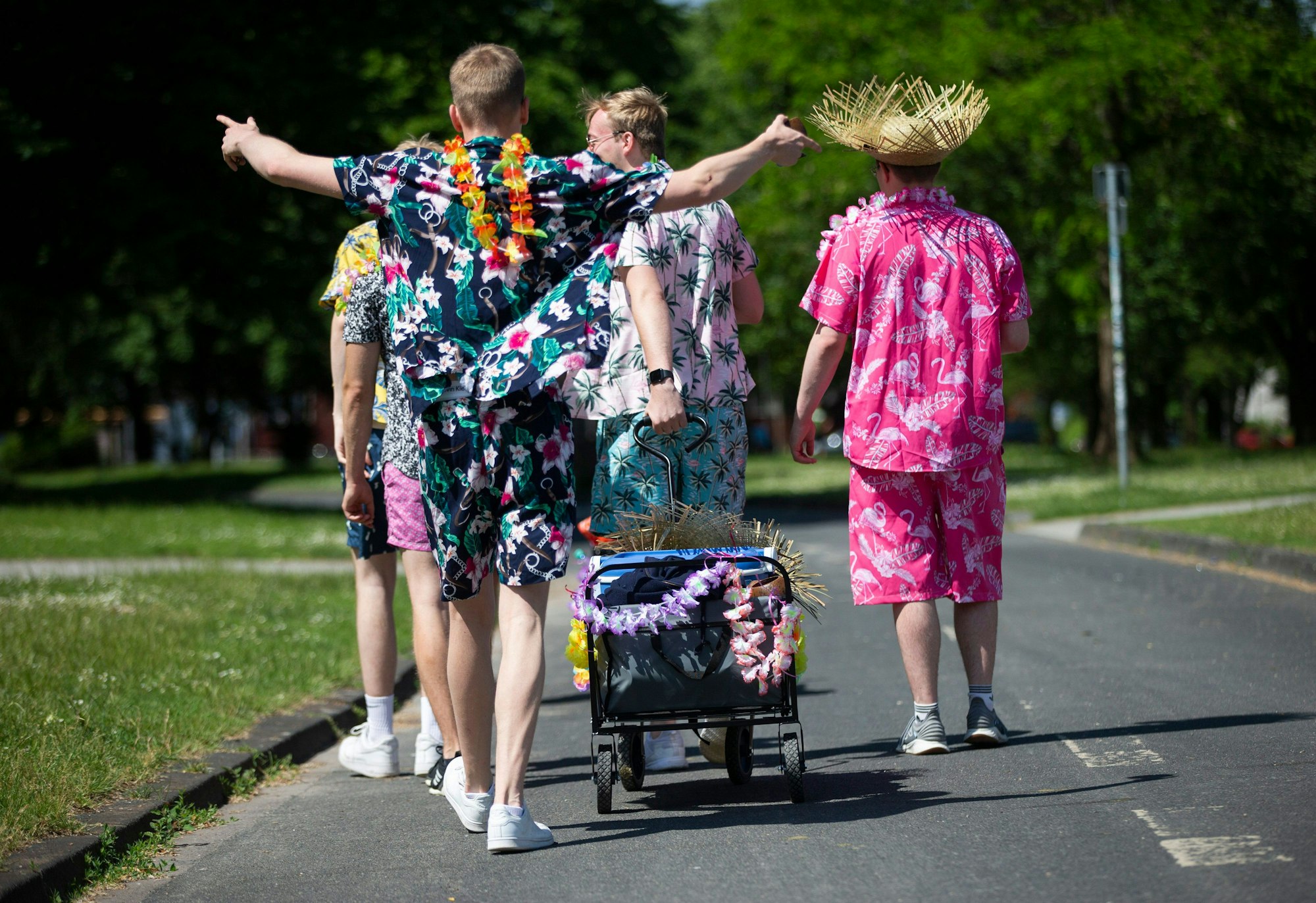 Junge Männer in Outfits mit floralen Mustern gekleidet, ziehen einen geschmückten Bollerwagen mit Getränken am Aachener Weiher hinter sich her.