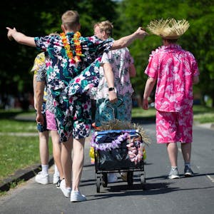 Junge Männer in Outfits mit floralen Mustern gekleidet, ziehen einen geschmückten Bollerwagen mit Getränken am Aachener Weiher hinter sich her.