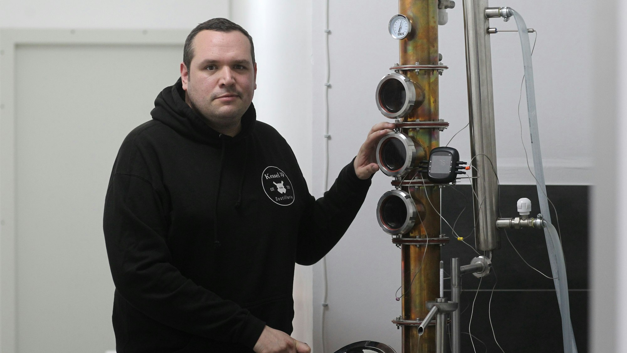 Kevin Braun aus Troisdorf schaut vor einem Gerät, das zur Gin-Produktion genutzt wird, in die Kamera. Er trägt einen schwarzen Hoodie.