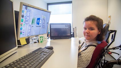 Julia Holler sitzt in einem Rollstuhl direkt vor ihrem Schreibtisch. Auf dem Schreibtisch stehen drei Bildschirme.
