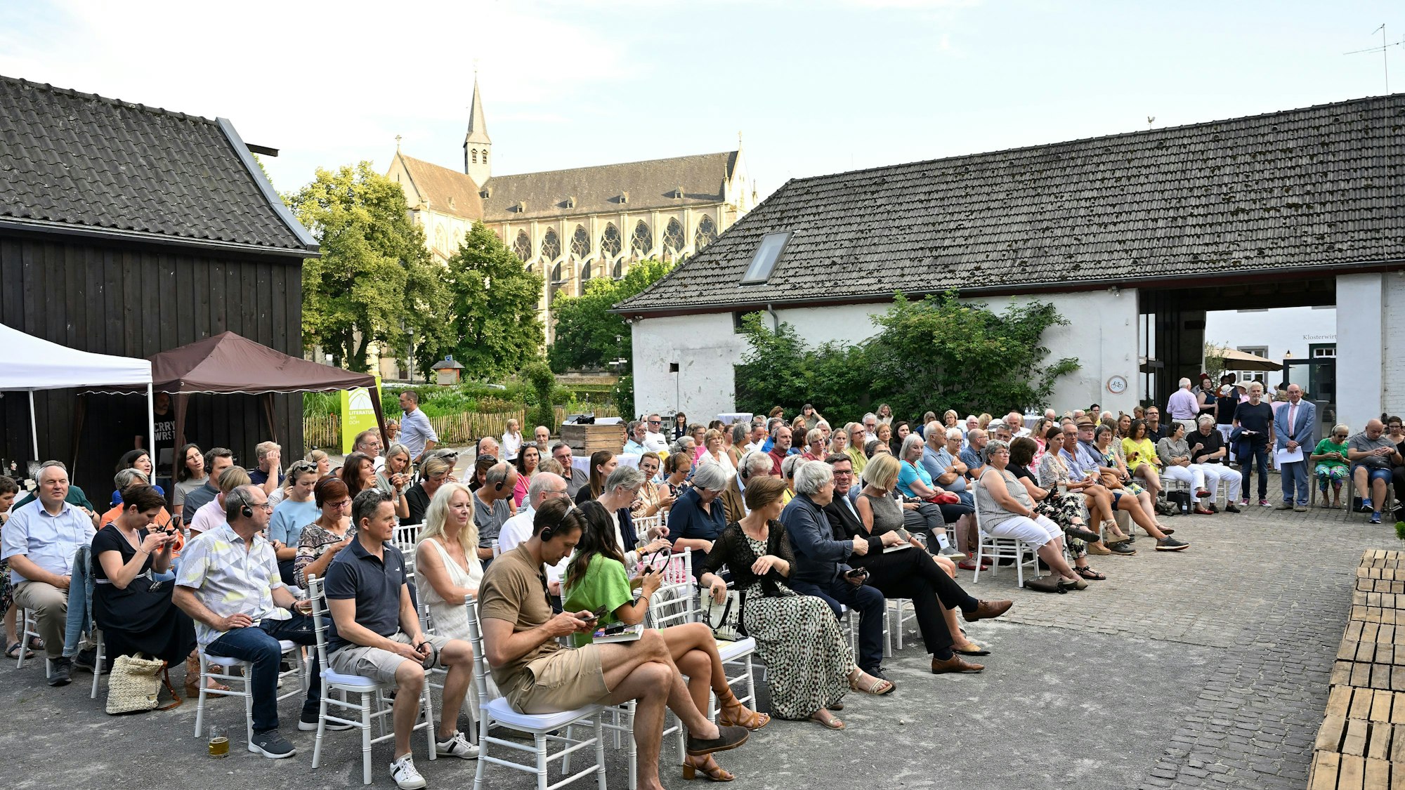 In einem Innenhof sitzen viele Menschen und blicken auf eine Bühne. Im Hintergrund ist der Altenberger Dom zu sehen.
