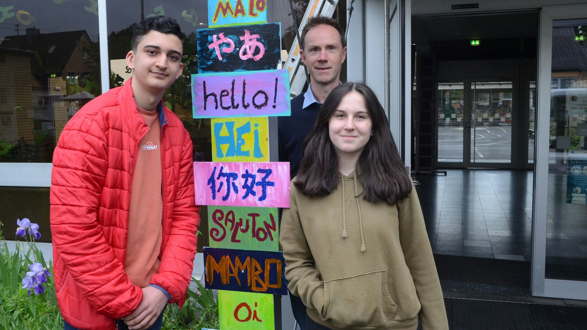 Von links: Schülersprecher Arman, Schulleiter Florian Heinick und Schülersprecherin Vivien stehen vor einem Begrüßungsschild, das Begrüßungen in verschiedenen Sprachen darstellt.