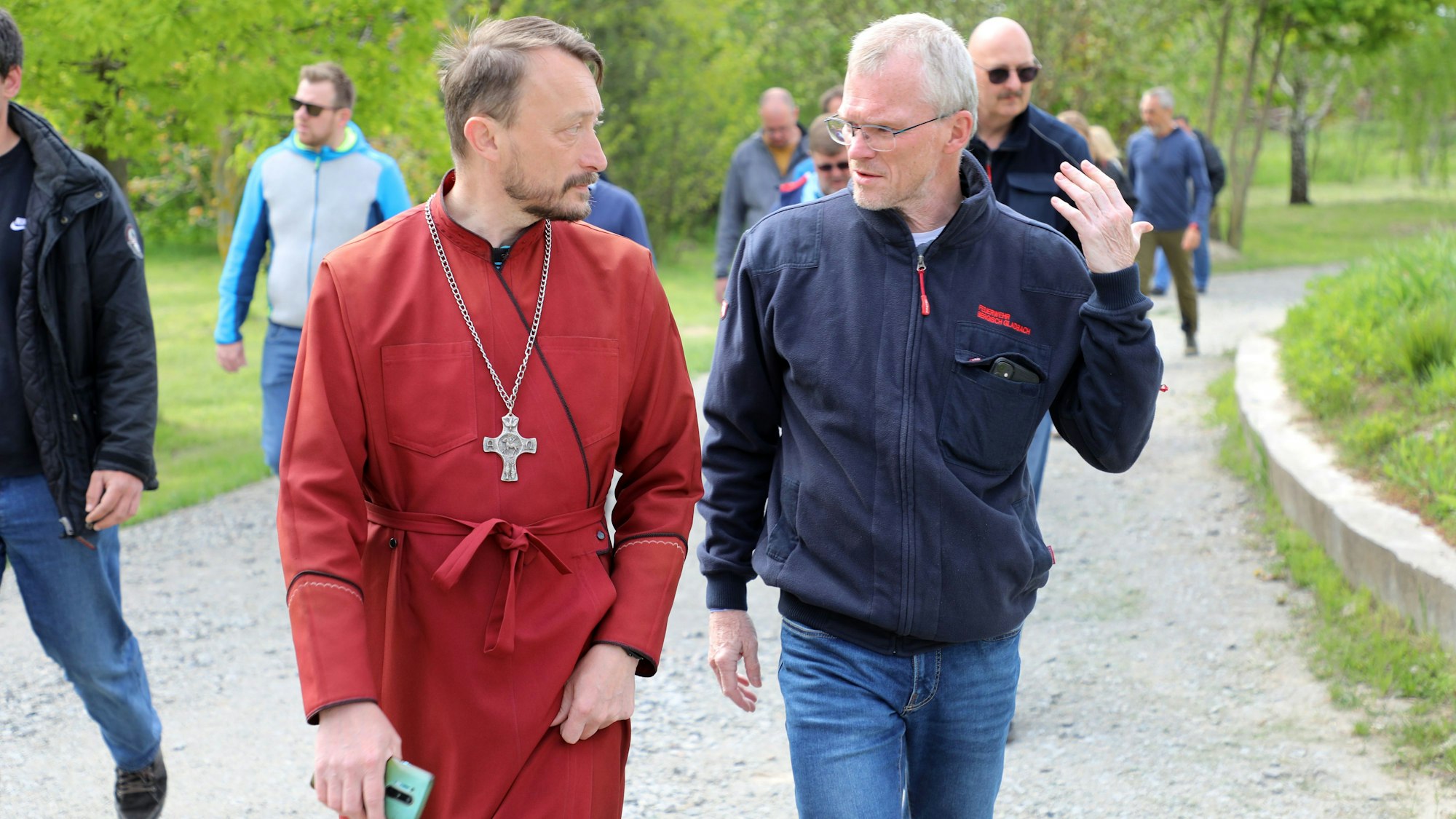 Ein orthodoxer Geistlicher und ein Mann mit Jacke der Feuerwehr Bergisch Gladbach unterhalten sich im Gehen.