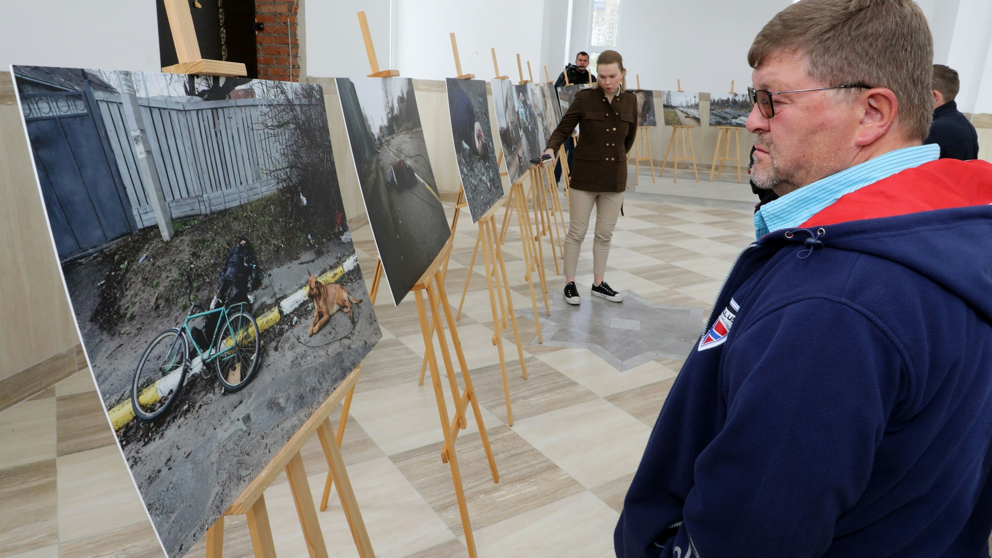 Ein Mann betrachtet ein Ausstellungsbild, auf dem ein erschossener Fahrradfaher in Butscha zu sehen ist, neben dem ein Hund auf der Straße sitzt.