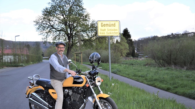Wolfgang Meyer aus Gemünd sitzt vor dem Ortsschild Gemünd auf seinem Motorrad und schaut in Richtung Kamera.