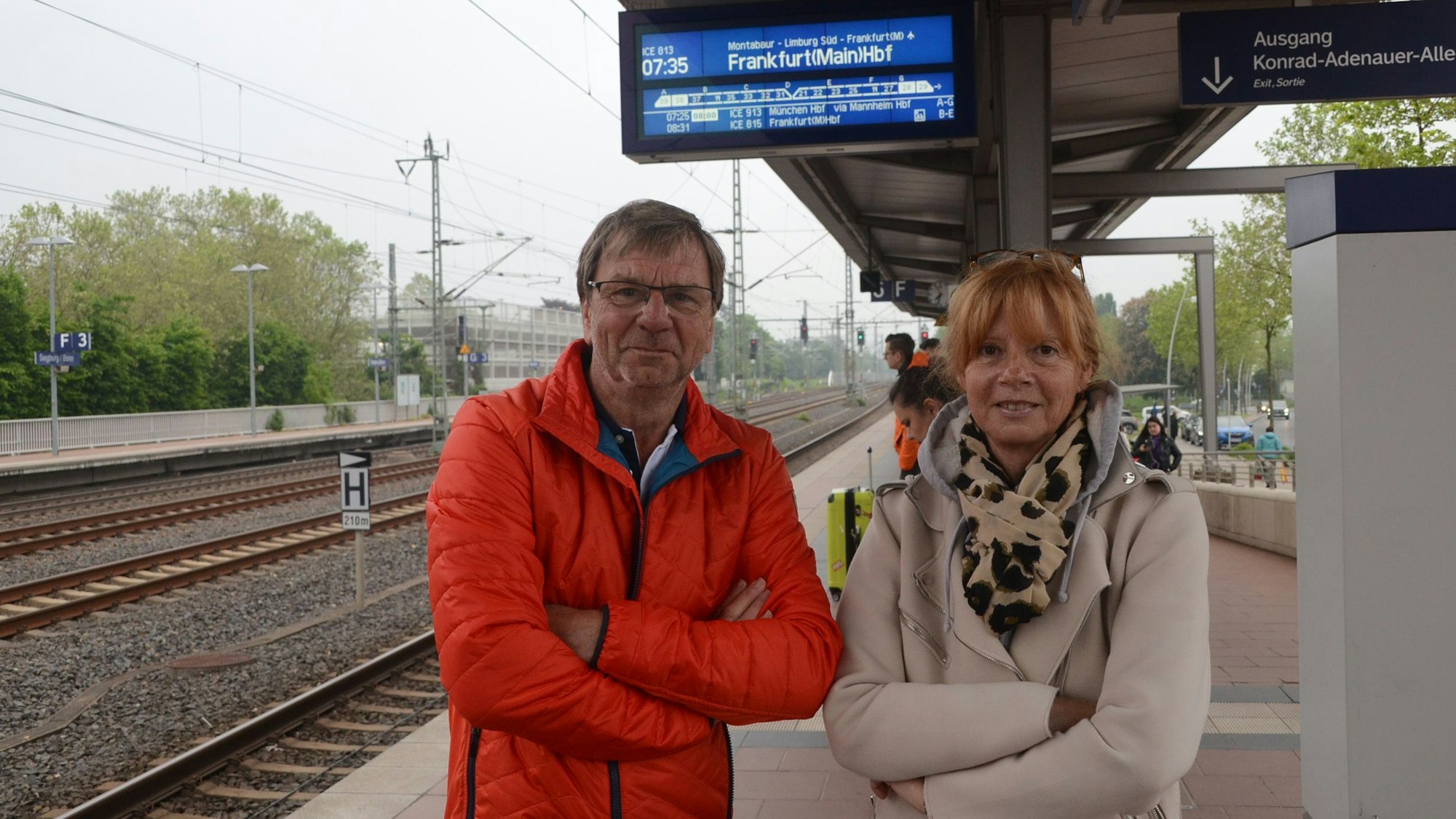 Ein Mann und eine Frau stehen auf dem Bahnsteig, neben ihnen liegen die Gleise, über ihnen hängt eine Anzeigetafel, die einen Zug nach Frankfurt ankündigt.