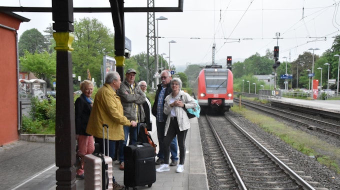 Auf einem Bahnsteig steht eine Gruppe Menschen mit Koffern, ein Zug fährt ein.