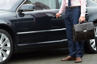 Vor einer schwarzen Limousine steht ein Mann in Anzughose, Hemd und Schlips, der eine Aktentasche trägt. Oberkörper und Kopf des Mannes sind nicht zu sehen. Es handelt sich um ein Symbolbild zum Thema Firmenwagen.