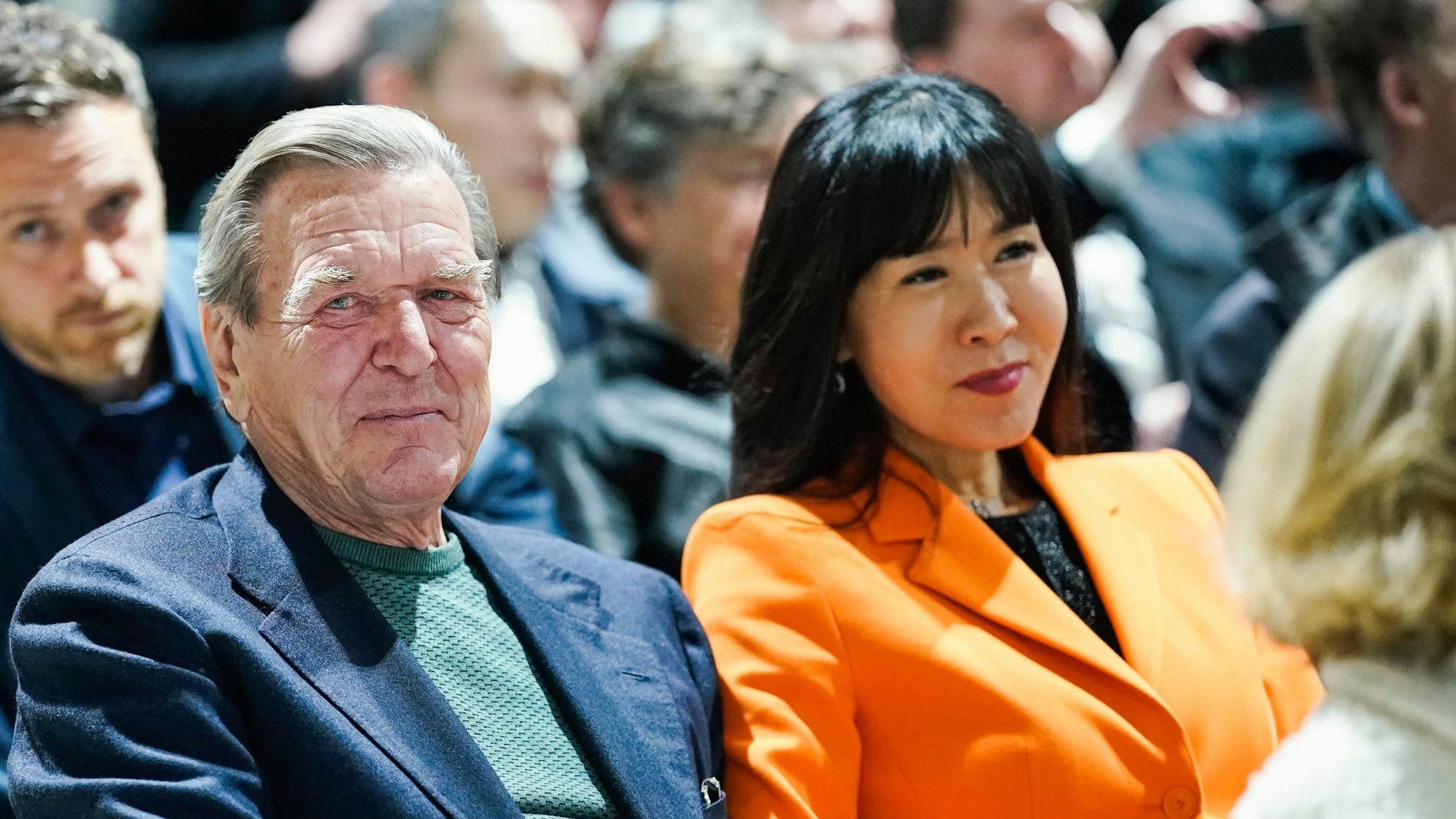 Der ehemalige Bundeskanzler Gerhard Schröder und seine Frau So-yeon Schröder-Kim sitzen im Publikum bei einer Veranstaltung. Frau Schröder-Kim trägt einen orangefarbenen Blazer.