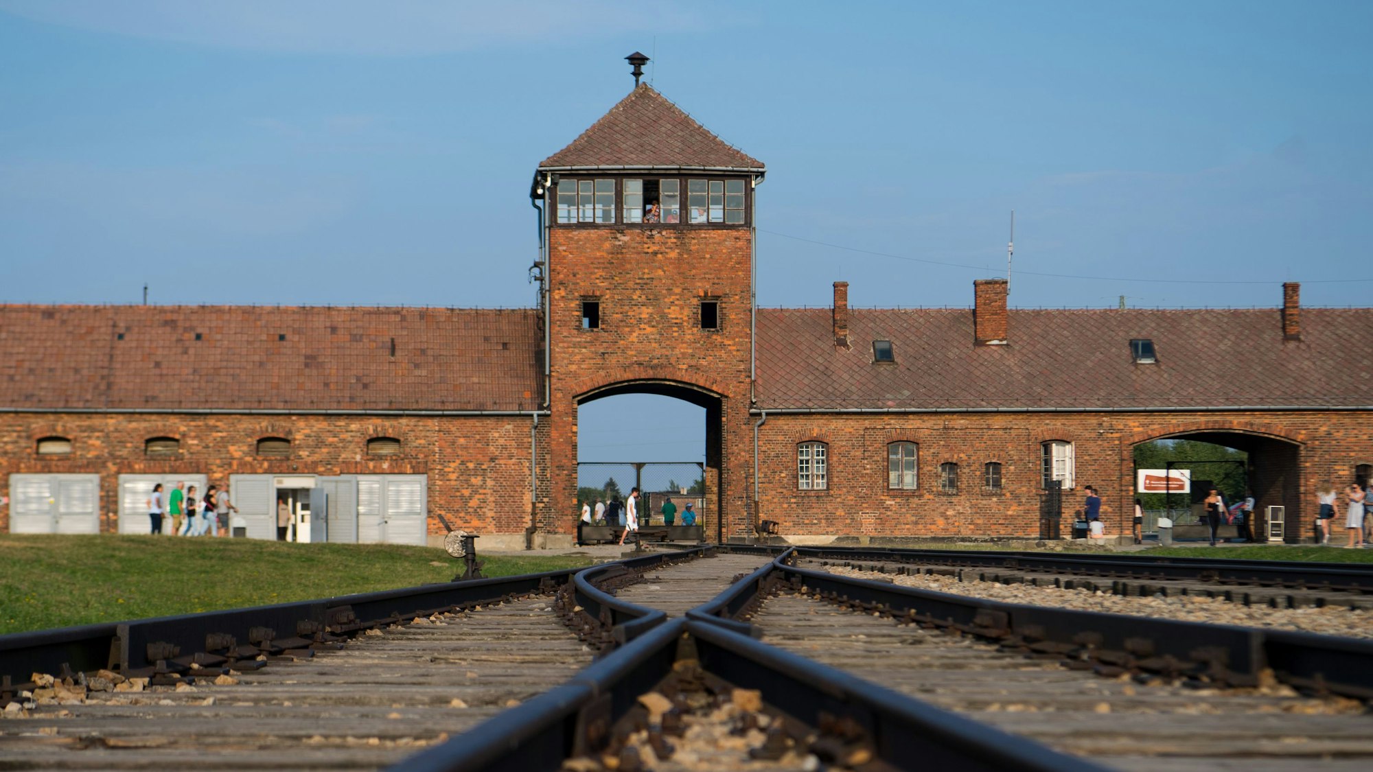 Besucher gehen am historischen Eingangstor des Vernichtungs- und Konzentrationslagers Auschwitz-Birkenau vorbei. Im Vordergrund sind die Gleise zu sehen, die in das Lager führen.