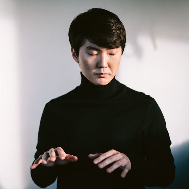 Pianist Seong-Jin Cho trägt einen schwarzen Rollkragen-Pullover und hält die Hände in die Luft, als würde er Klavier spielen.
