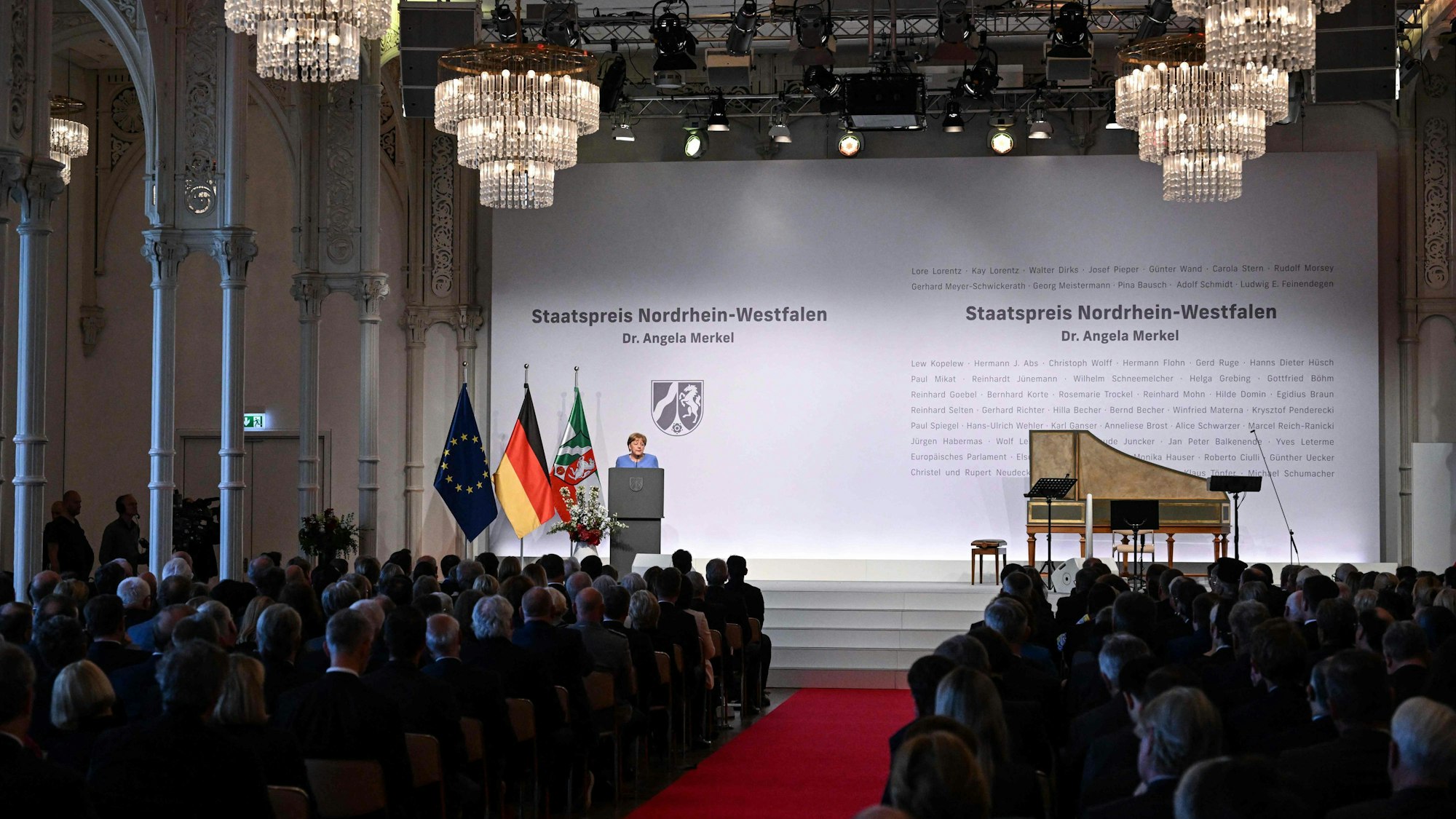 Angela Merkel spricht auf der Bühne in der Kölner Flora. Sie ist von weit hinten im Saal fotografiert, man sieht Publikum und einen Flügel neben ihr auf der Bühne. Ebenso die Flaggen von Europa, Deutschland und NRW.