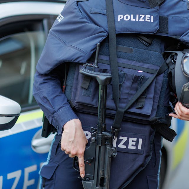 Eine Polizistin steht mit ballistischem Schutzhelm, einer Schusswaffe und einer kugelsicheren Weste vor einem Polizeiwagen.