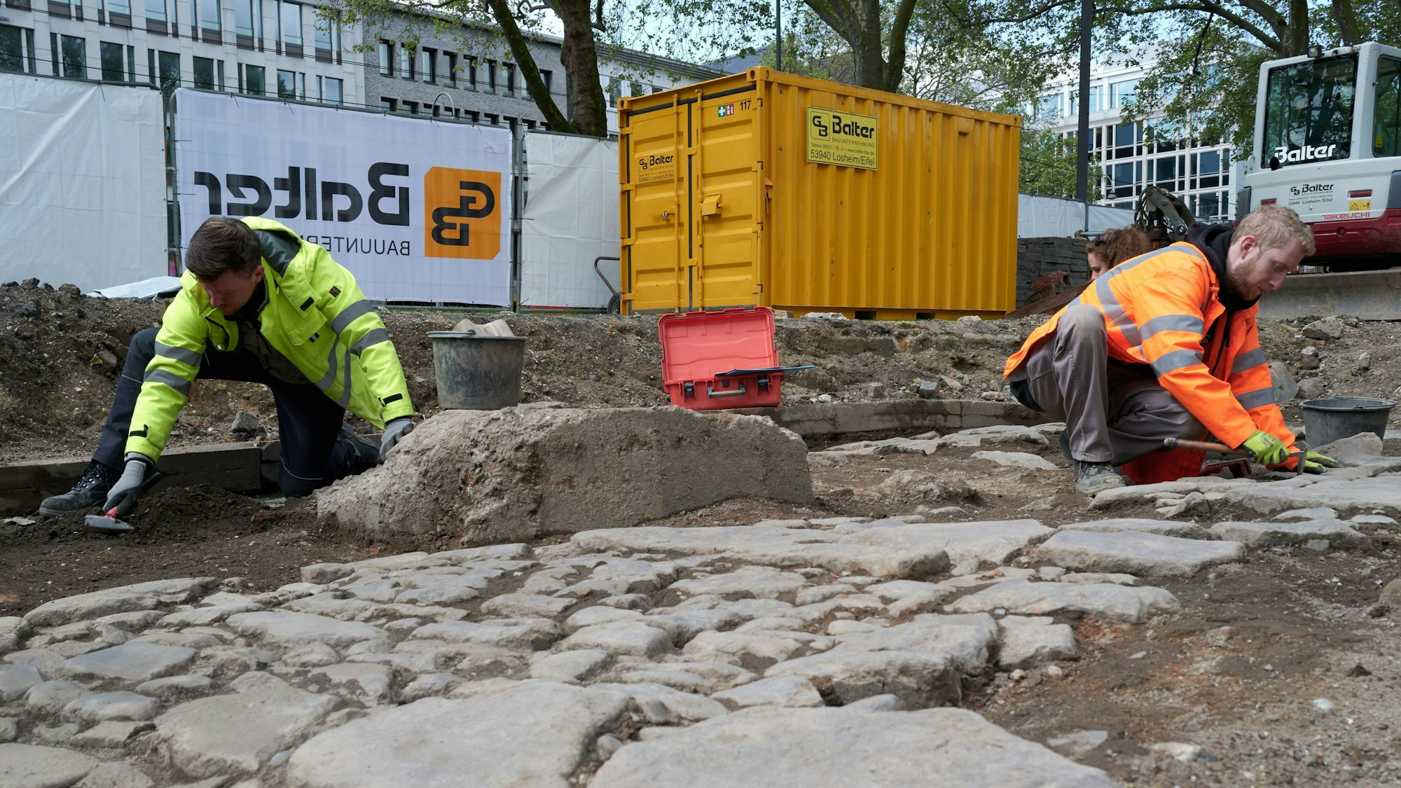 Zwei Bauarbeiter, die eine abgesperrte Fläche mit Steinen bearbeiten