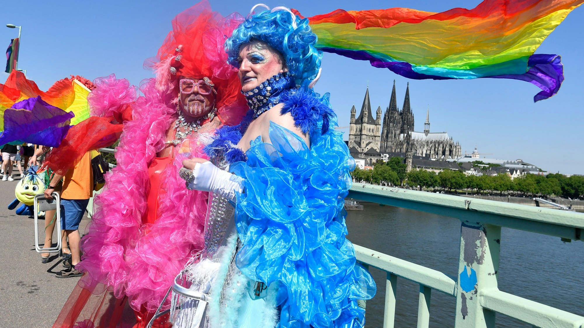 Beim CSD in Köln hatten sich viele Teilnehmer fantasievoll verkleidet. Zwei Teilnehmer stehen in knallfarbenen Roben am Rheinufer, im Hintergrund weht eine Regenbogenfahne.
