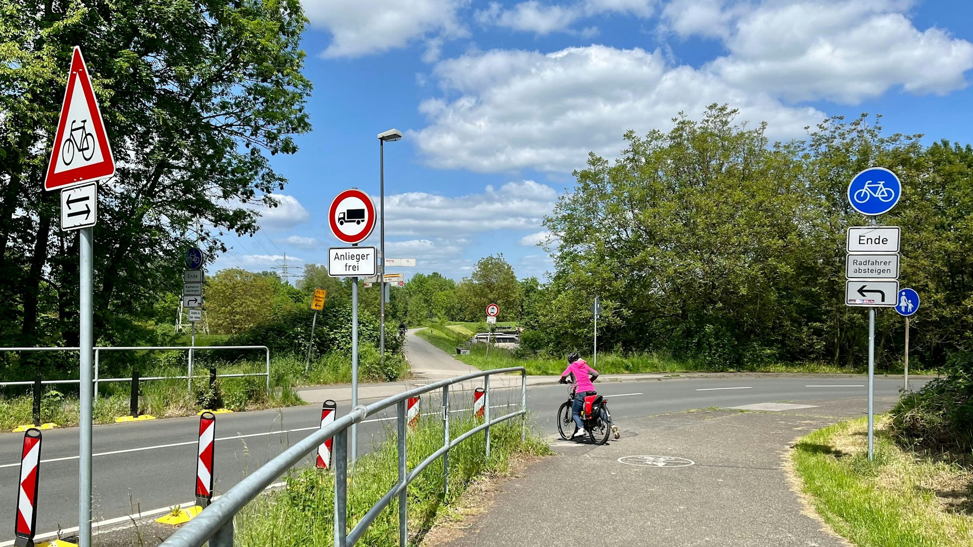 An der Einmündung der Rheindorfer Straße in den Westring widersprechen sich die Verkehrsschilder. Eine Radlerin wagt es, nicht abzusteigen.