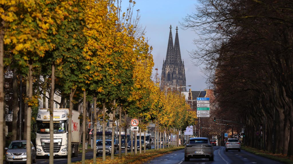Auto fährt auf einer Allee in Köln entlang, im Hintergrund ist der Kölner Dom zu sehen.