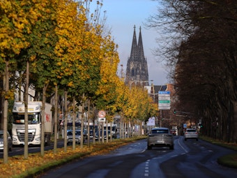 Auto fährt auf einer Allee in Köln entlang, im Hintergrund ist der Kölner Dom zu sehen.